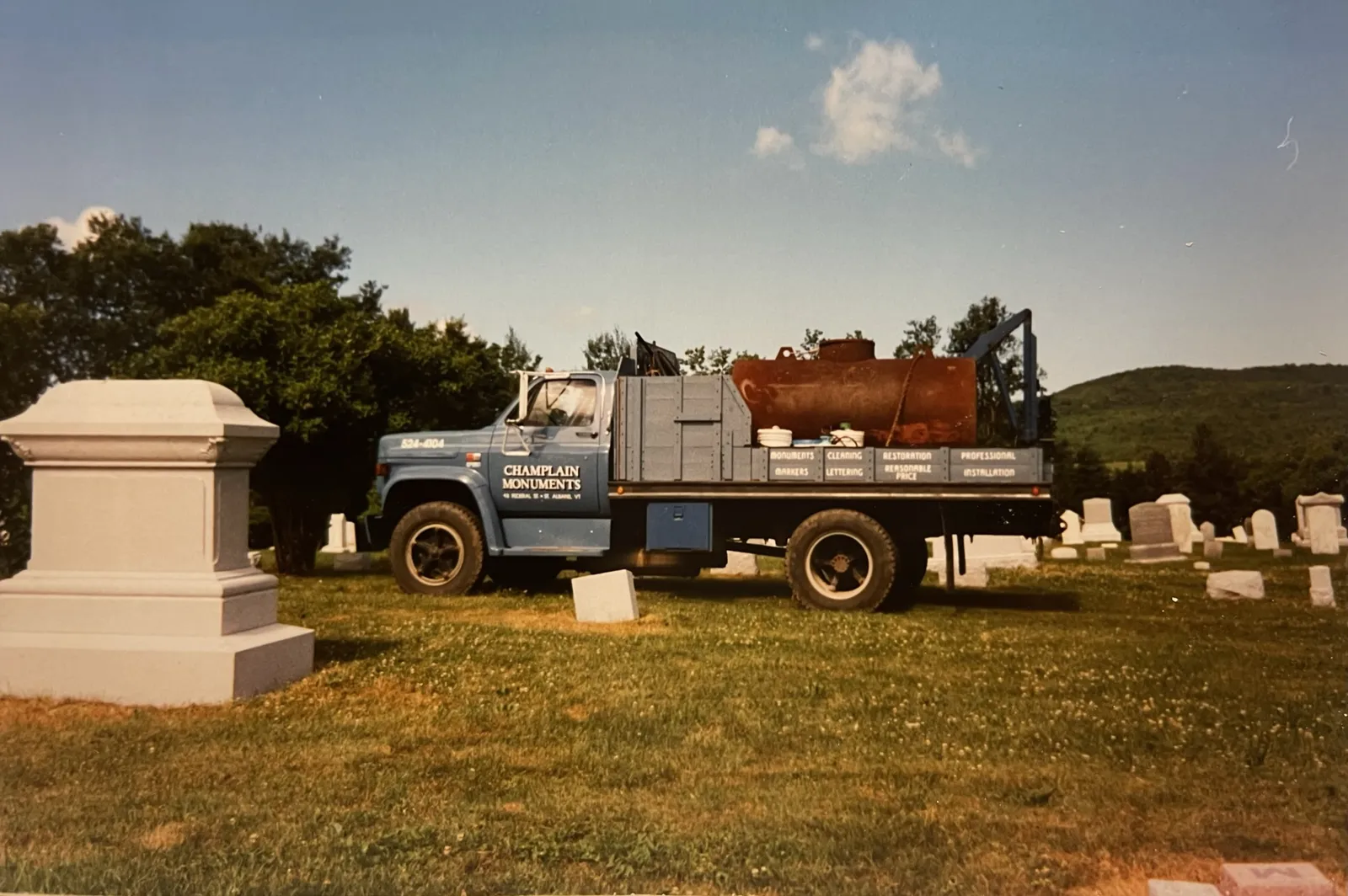 The original Champlain Monuments truck at a cemetery job, with services painted on the side panel — monuments, markers, cleaning, lettering, restoration, professional installation