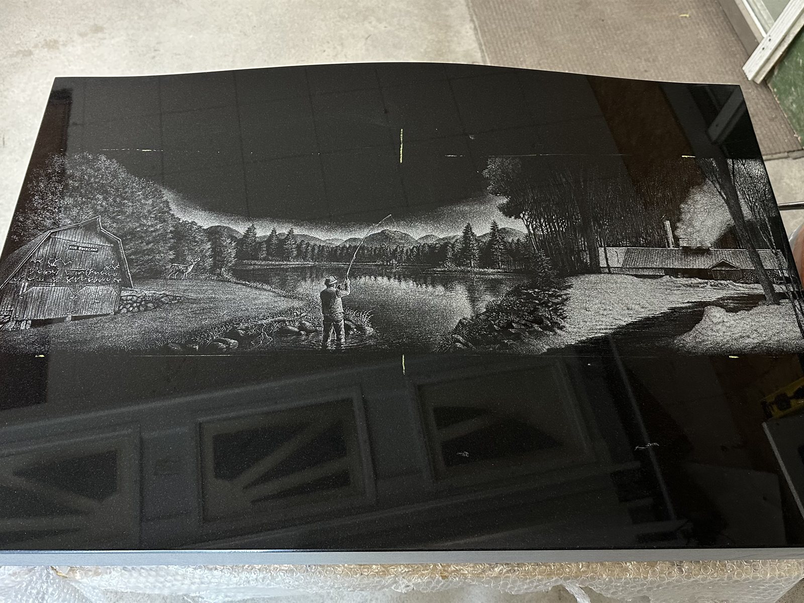 The completed hand-etched scene on polished black granite — a fly fisherman on a pond with mountains, a labeled Five Buck Camp barn, and a sugar shack with smoke from the chimney
