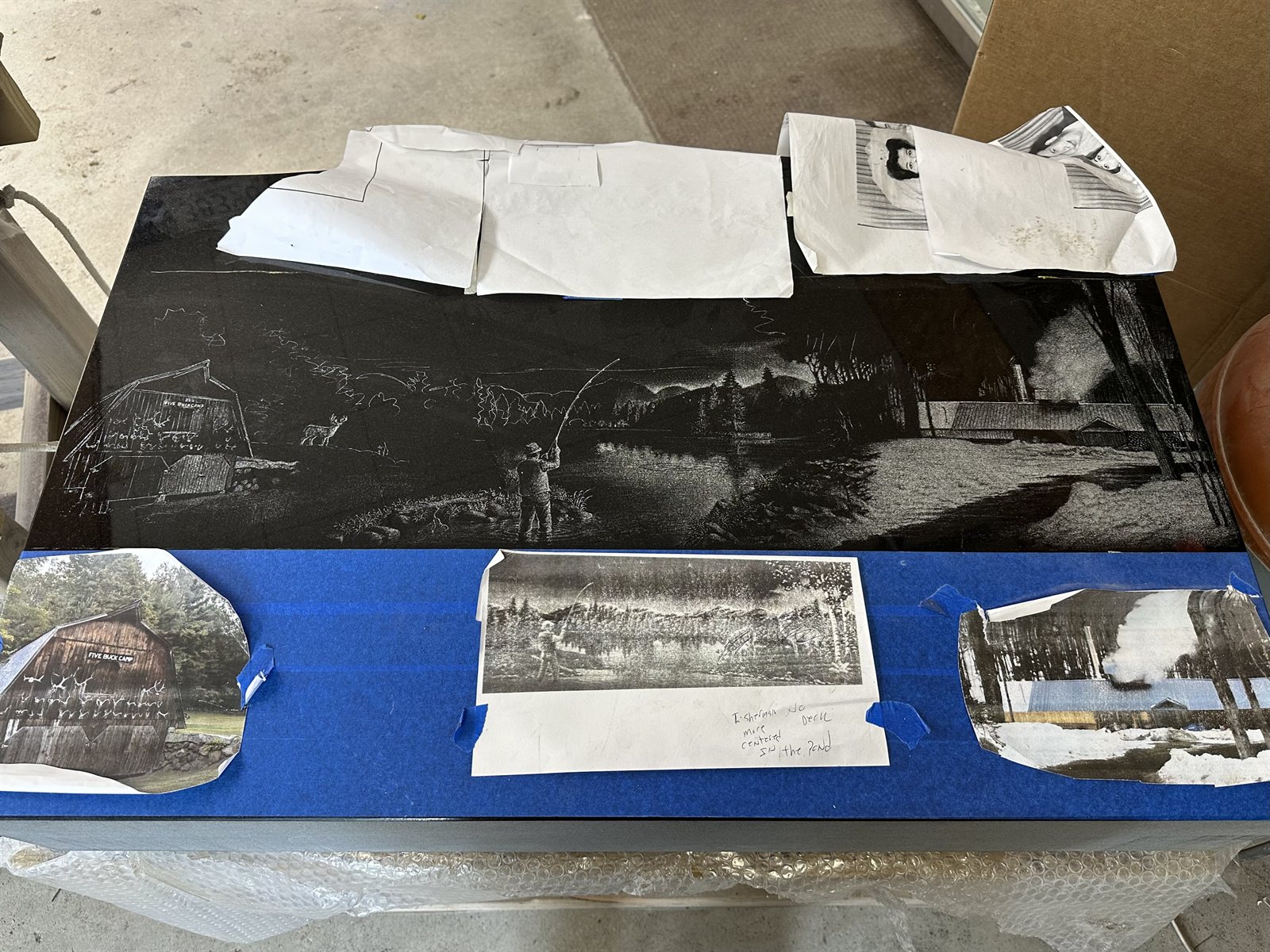 The same granite slab partially hand-etched, with the lower half of the scene roughed in and the original sketches still taped above