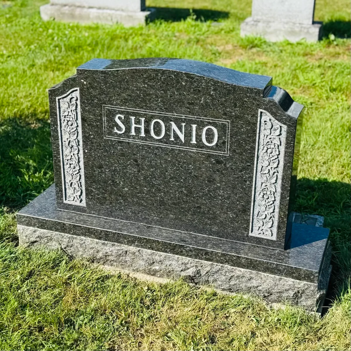 The Shonio memorial — dark green polished upright with hand-carved floral border panels and a raised name panel